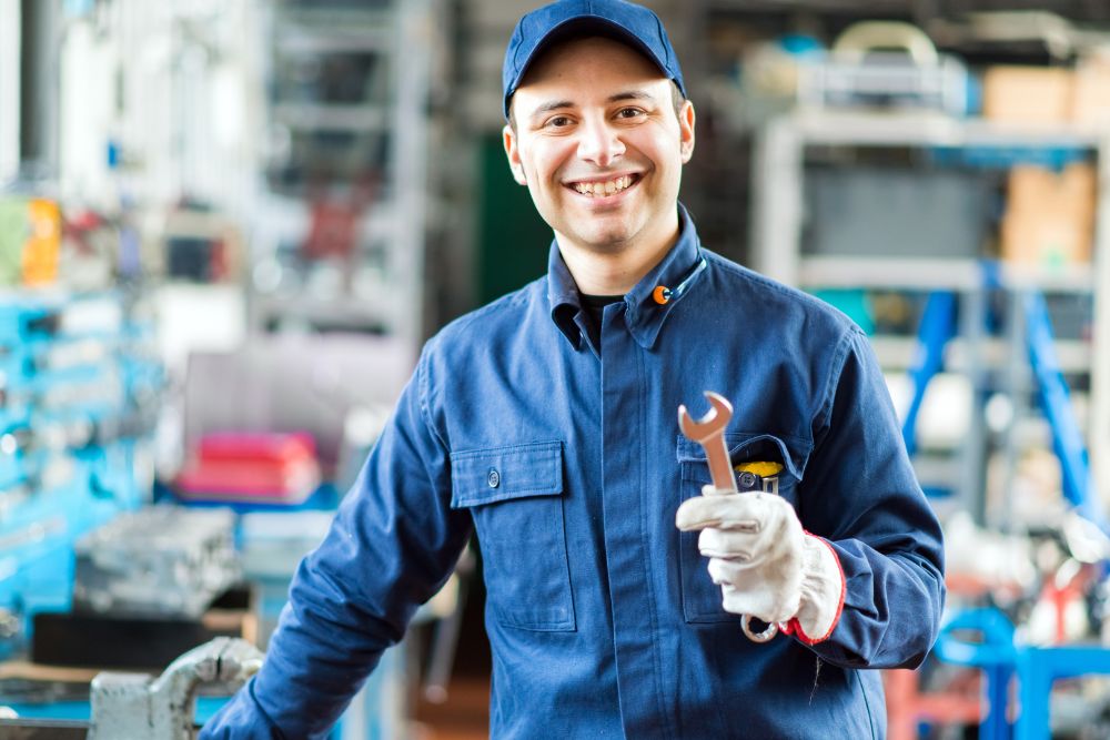 Automotive technician smiling while holding a wrench in an auto repair shop, emphasizing the importance of vehicle maintenance and tune-ups.